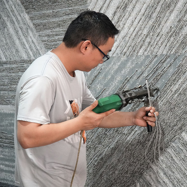 A hand tufting craftsman is working on a rug with grey abstract patterns, wearing a cream t-shirt