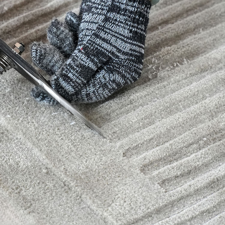 A custom tufted rug craftsman carves the high piles of a beige wool rug to create texture variations