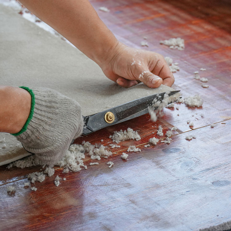 A tufting worker holds a corner of a beige tufted rug with one hand, and trim the edge with scissors using another hand