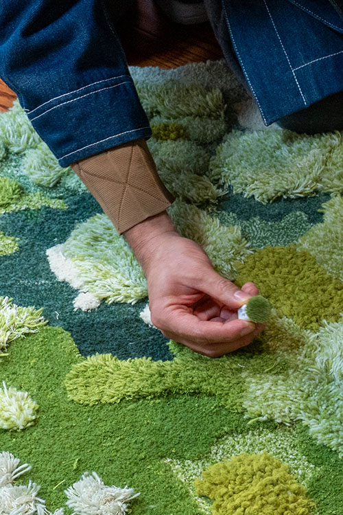 Custom rug tufting artisan holding soft yarn samples on a green woolen moss rug with varied pile heights