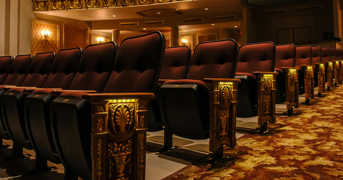 A theater with rows of plush burgundy-colored seats that have intricately gold-detailed aisle ends. The seats are on a patterned, wall-to-wall carpet in rich red and gold.