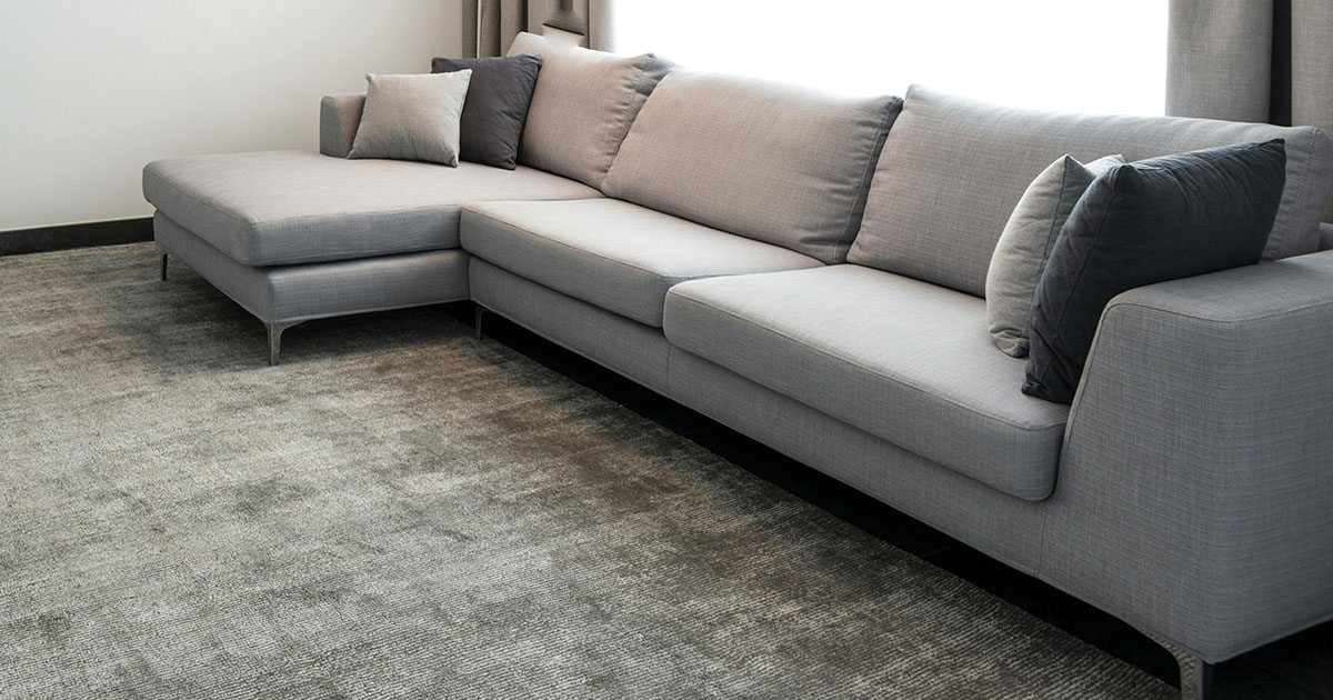 Modern living room with a light gray L-shaped sectional sofa and dark and light gray accent pillows. On the floor is a low-pile custom area rug in silver-gray hues that complements the minimalist décor.