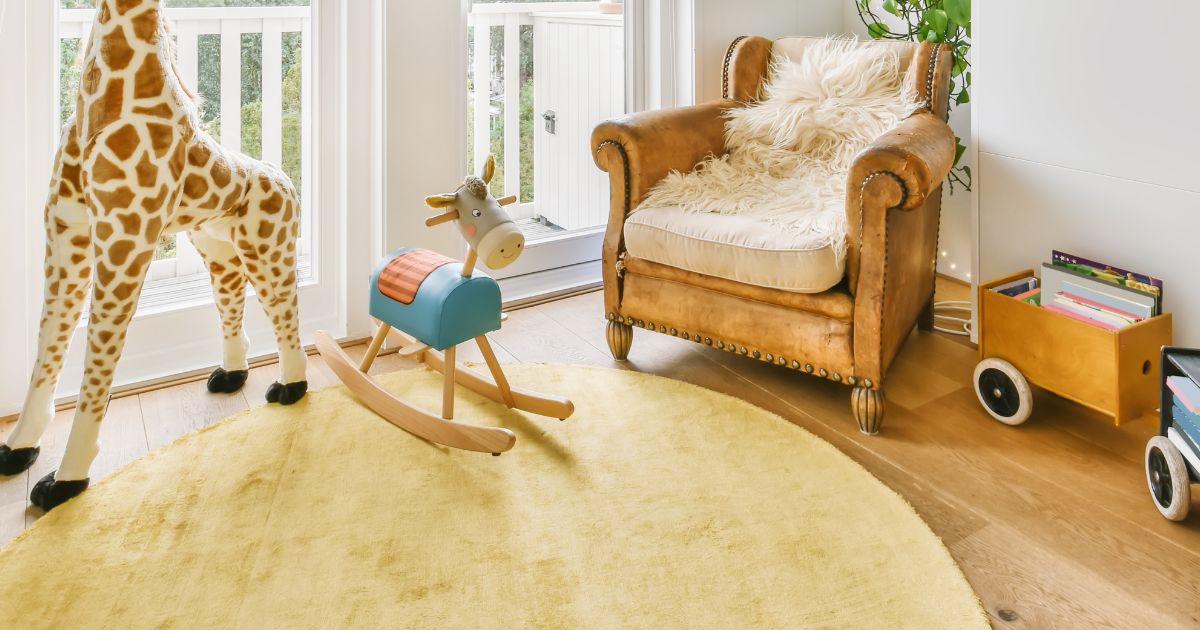 Vibrant kids play area with the white round pale yellow area rug over light wood floors for a cozy nursery corner. The space includes leather armchair with sheepskin throw, large decorative giraffe stuffed toy.